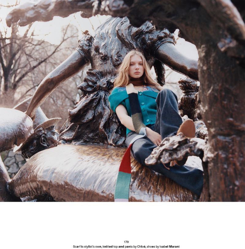 Model Secret Snow sits on a bronze park sculpture wearing a teal knit top, multi-color scarf, and dark trousers.