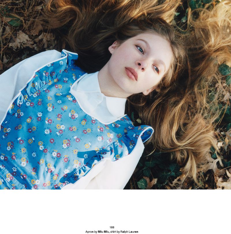 Model Secret Snow lying in dry foliage wearing a blue floral pinafore dress over a white collared shirt.