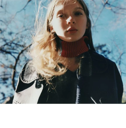 Model Secret Snow wearing a dark coat and red knit collar, looking down towards the camera against bare trees and blue sky.