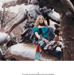 Model Secret Snow sits on a bronze park sculpture wearing a teal knit top, multi-color scarf, and dark trousers.