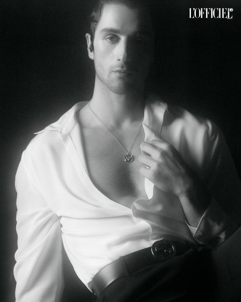 Agustín Della Corte posing in an unbuttoned white shirt and pendant necklace against a dark background.