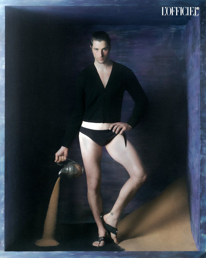 Man wearing a black cardigan and briefs holding a pitcher pouring sand inside a dark textured enclosure for L'Officiel Chile