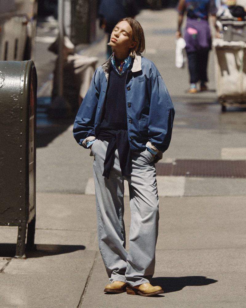 Lena Mantler standing in direct sunlight on a city sidewalk wearing a blue collared jacket, grey trousers, and tan boots.