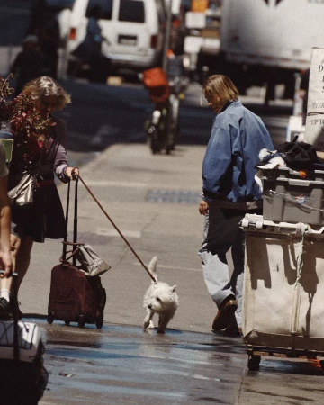 Model Lena Mantler in a blue jacket walking down an urban city street sidewalk next to rolling carts and a small white dog.