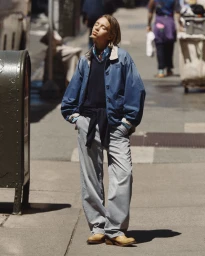 Lena Mantler standing in direct sunlight on a city sidewalk wearing a blue collared jacket, grey trousers, and tan boots.