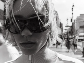 Black and white close-up portrait of Lena Mantler wearing dark wraparound sunglasses and a ribbed zip turtleneck on a city street.