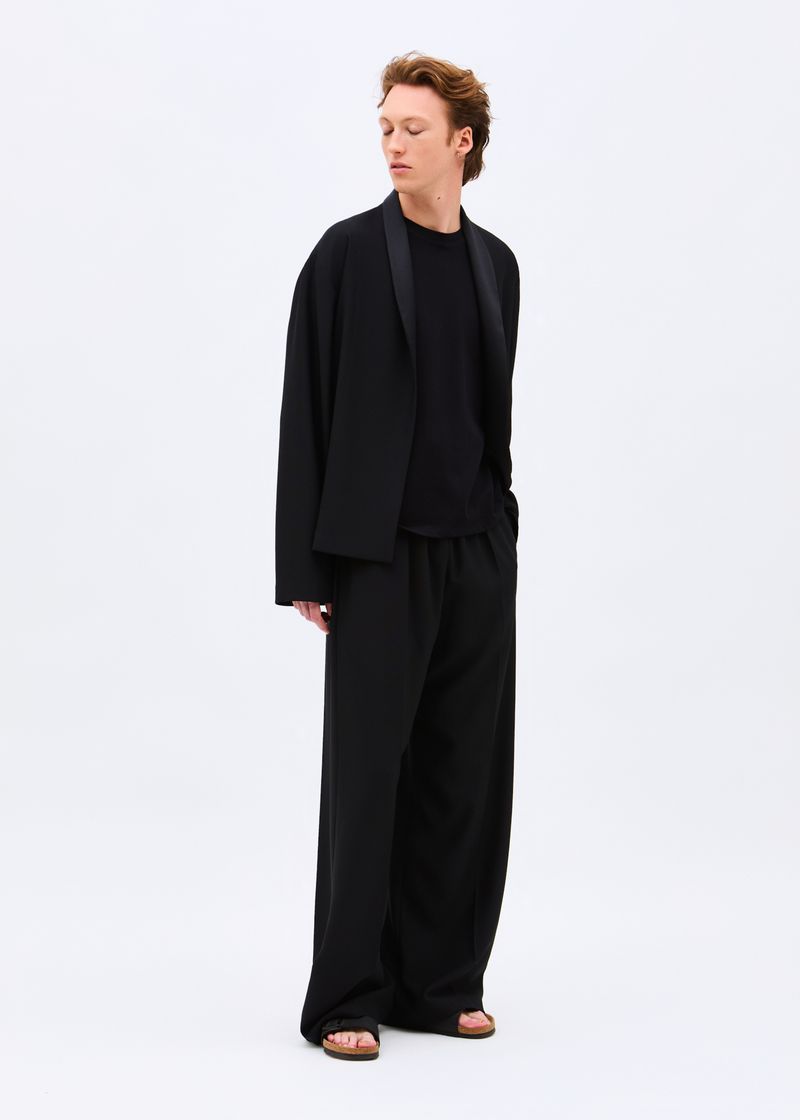 Model wearing an open black tailored jacket, wide-leg trousers, and sandals against a plain white studio backdrop.