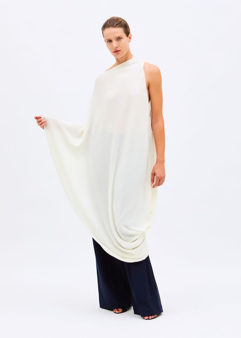 Model posing in an asymmetrical draped off-white tunic top with wide-leg navy trousers against a white seamless background.