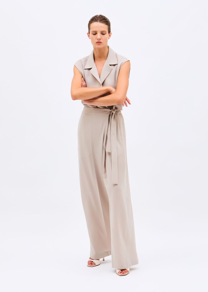 Model standing with arms crossed wearing a sleeveless beige collared jumpsuit with wide trousers against a white background.