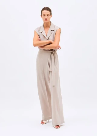 Model standing with arms crossed wearing a sleeveless beige collared jumpsuit with wide trousers against a white background.
