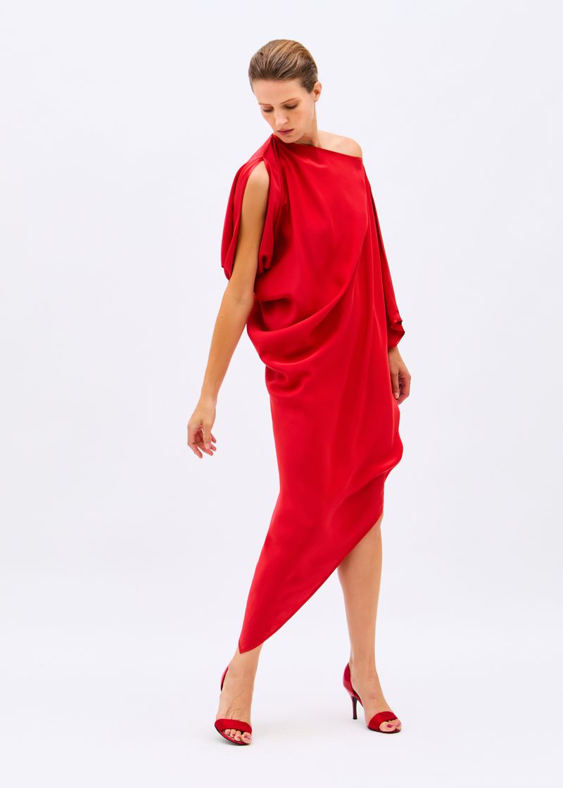 Model posing in an asymmetrical red draped dress and red heels against a seamless white studio background.