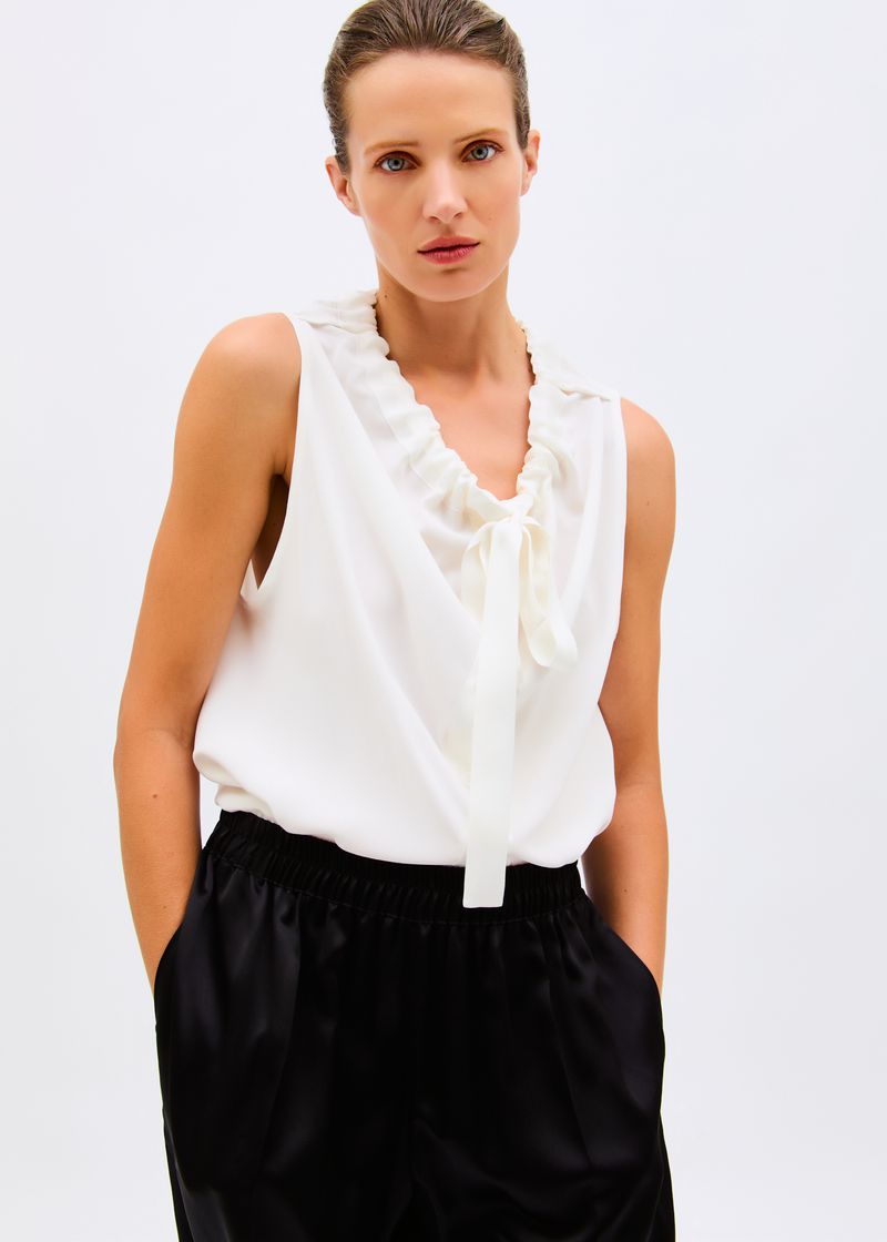 Model wearing a sleeveless white ruffled neck blouse and black satin pants against a seamless white studio background.