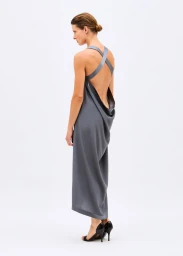 Model wearing a draped backless grey maxi dress with cross straps and black heels against a white seamless background.