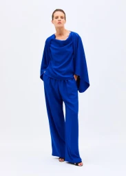 Model wearing a draped royal blue cape top and matching wide-leg trousers against a clean white seamless studio background.