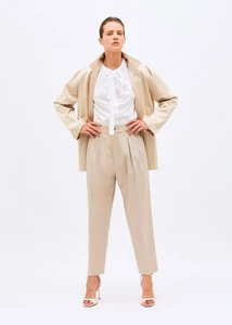 Model wearing a beige oversized blazer, matching pleated trousers, and a white tie-neck blouse against a white seamless backdrop.