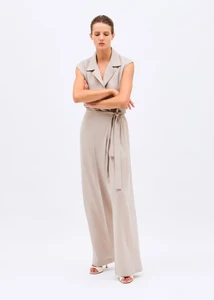 Model standing with arms crossed wearing a sleeveless beige collared jumpsuit with wide trousers against a white background.