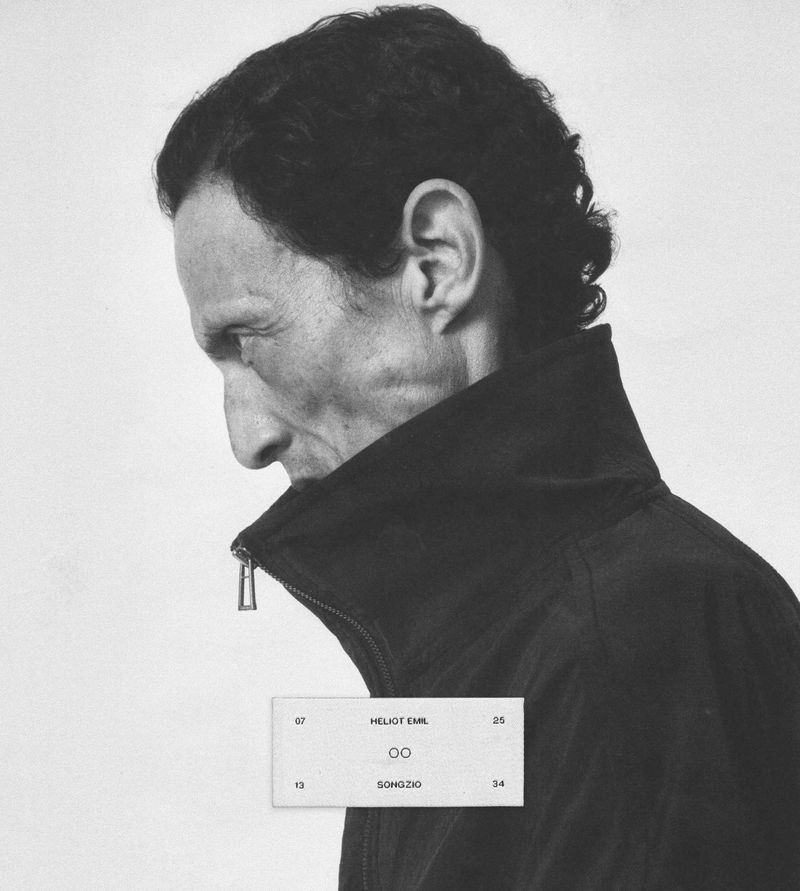 Black and white profile portrait of a man wearing a high-collar black zip jacket by Songzio and Heliot Emil against a plain background.