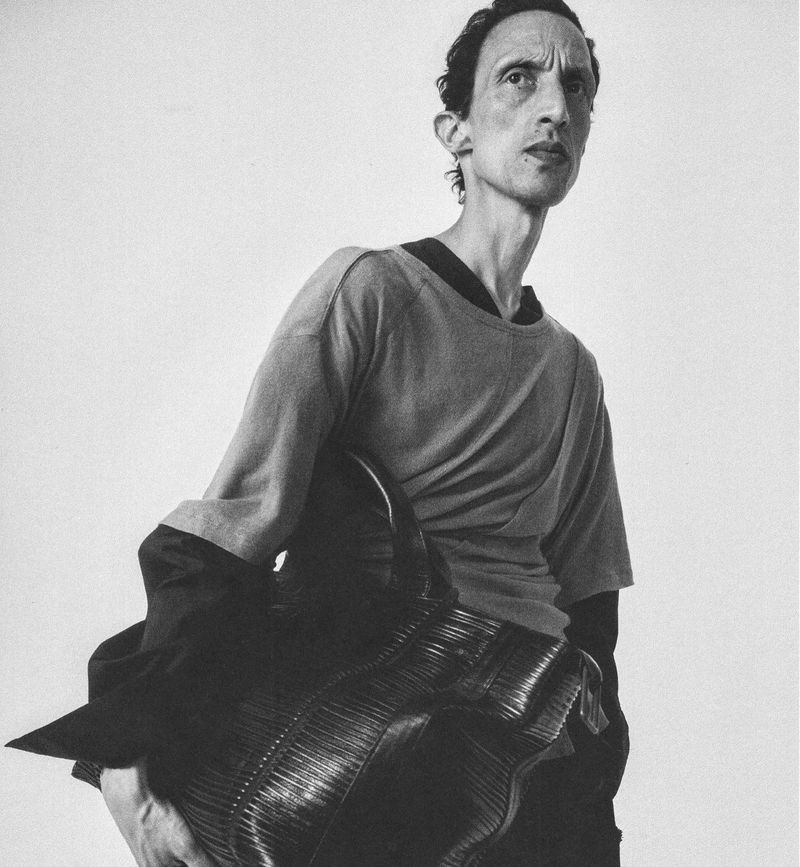 Monochrome portrait of a man holding a large textured dark leather tote bag against a plain white studio backdrop.