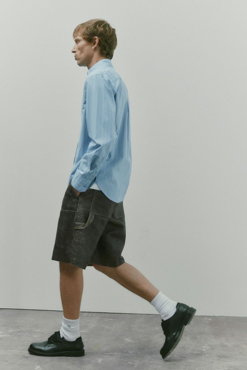 Model Mike Vasilakis walks in profile wearing a light blue button-down shirt, dark charcoal shorts, and black shoes in a studio setting.