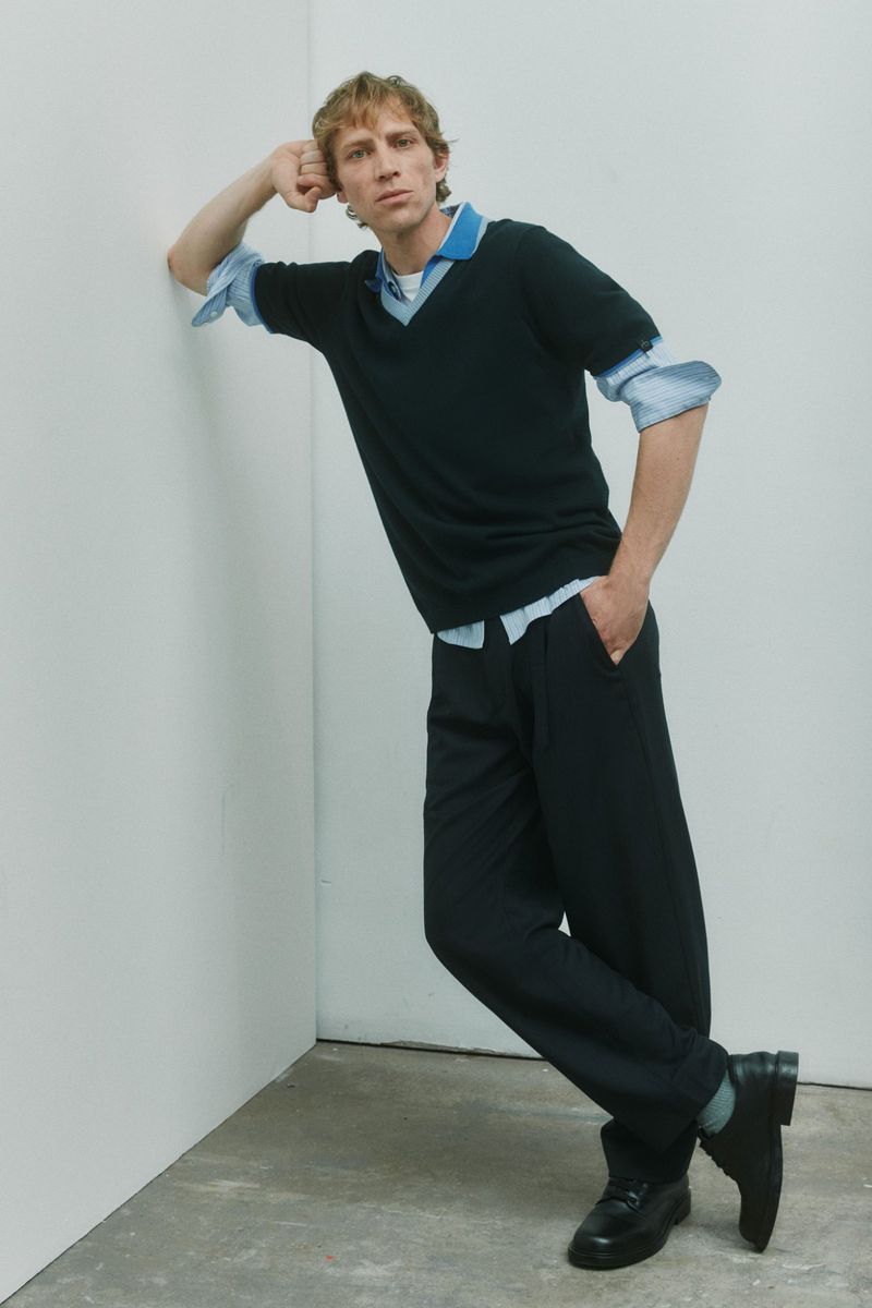 Model Mike Vasilakis leaning against a white studio wall wearing a black sweater over a blue collared shirt and black trousers.