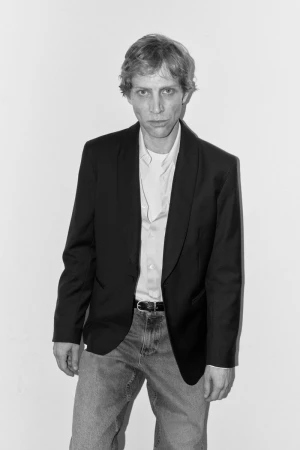 Model Mike Vasilakis in a black blazer, white collared shirt, and denim jeans against a white wall in black and white.
