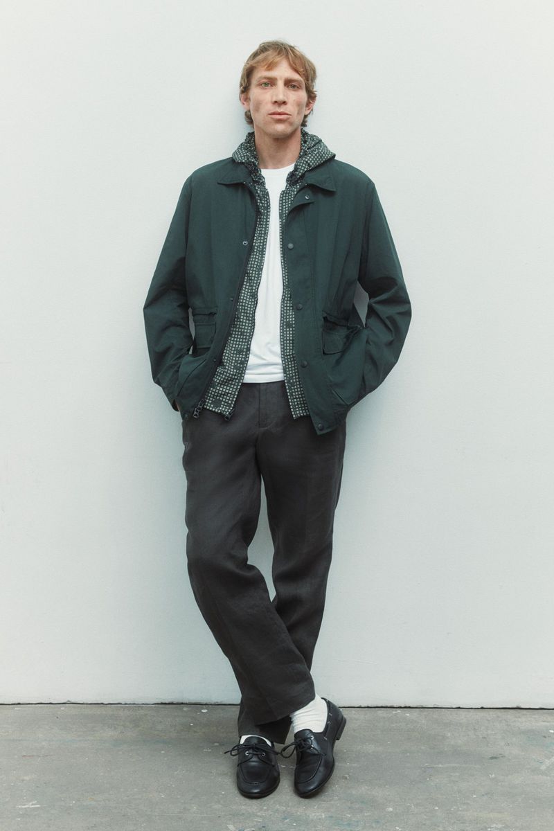 Model Mike Vasilakis standing against a white backdrop wearing a dark green jacket, grey trousers, and black loafers.