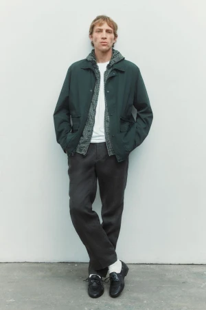 Model Mike Vasilakis standing against a white backdrop wearing a dark green jacket, grey trousers, and black loafers.