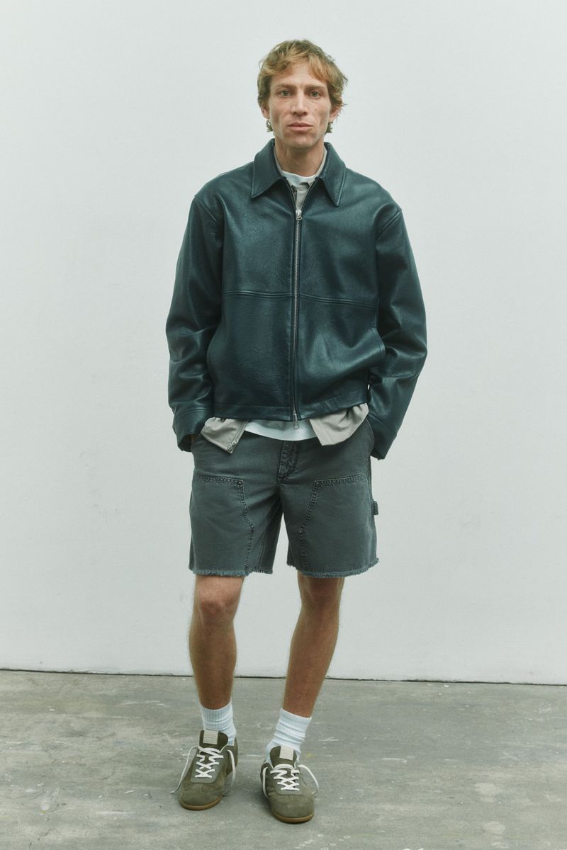 Model Mike Vasilakis wearing a dark leather zip jacket, grey denim shorts, and olive sneakers for Rag & Bone.