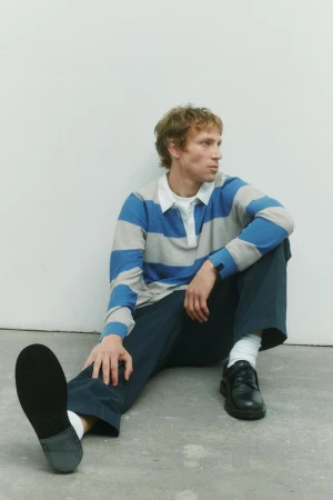 Model Mike Vasilakis seated on the floor wearing a blue and grey striped polo shirt and dark trousers for Rag & Bone.