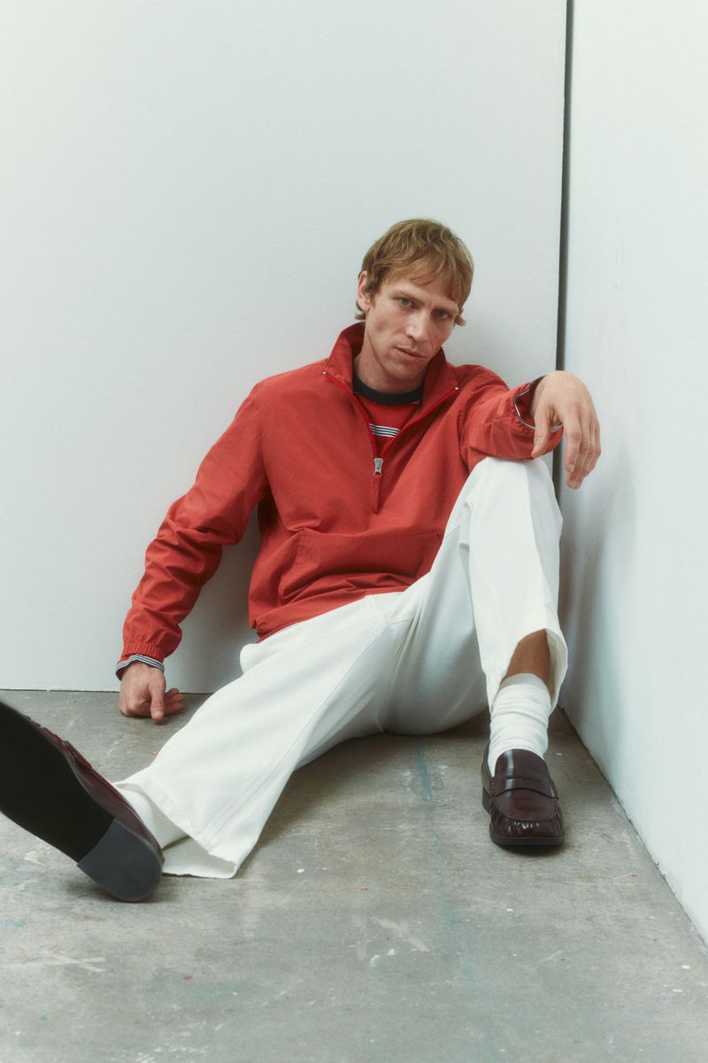 Model Mike Vasilakis seated on the floor wearing a red zip jacket, wide-leg white trousers, and dark loafers for Rag & Bone.