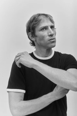 A man in a black ringer t-shirt with white trim posing against a seamless light grey studio backdrop in black and white.