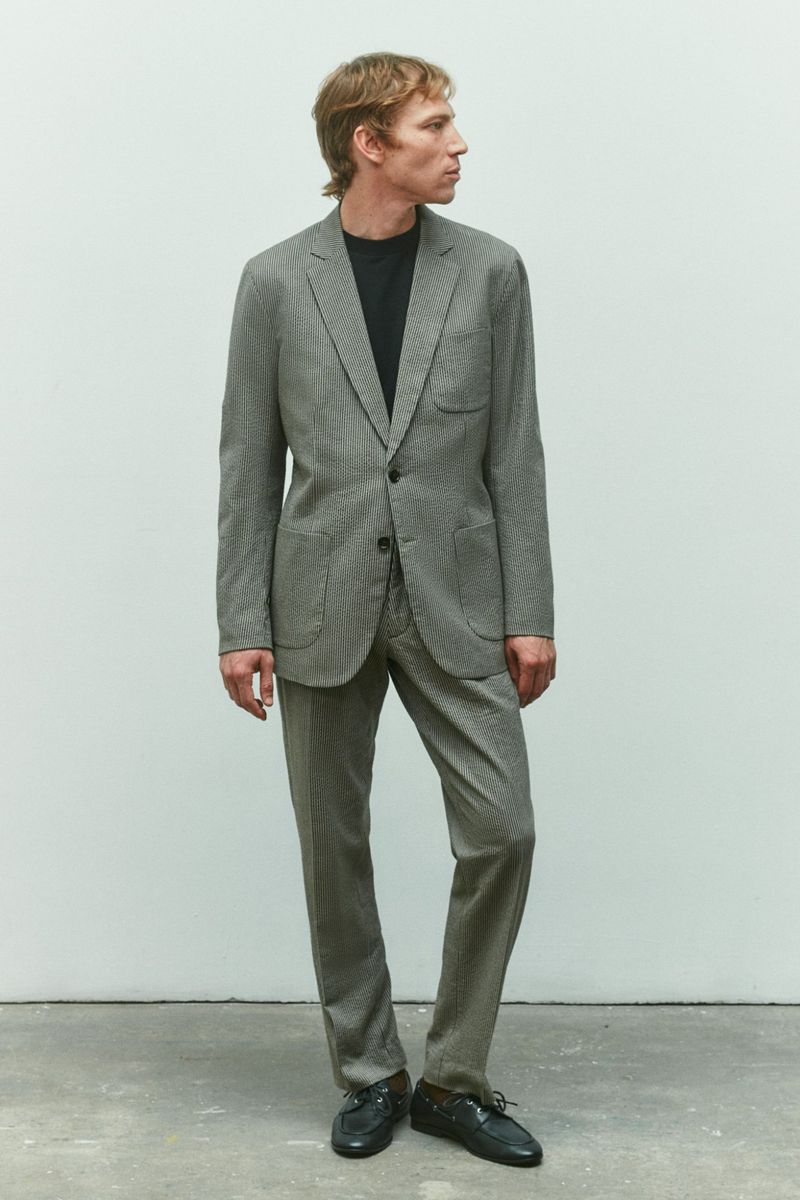 Model Mike Vasilakis wearing a grey tailored suit and black crewneck shirt against a neutral studio backdrop for Rag & Bone menswear.