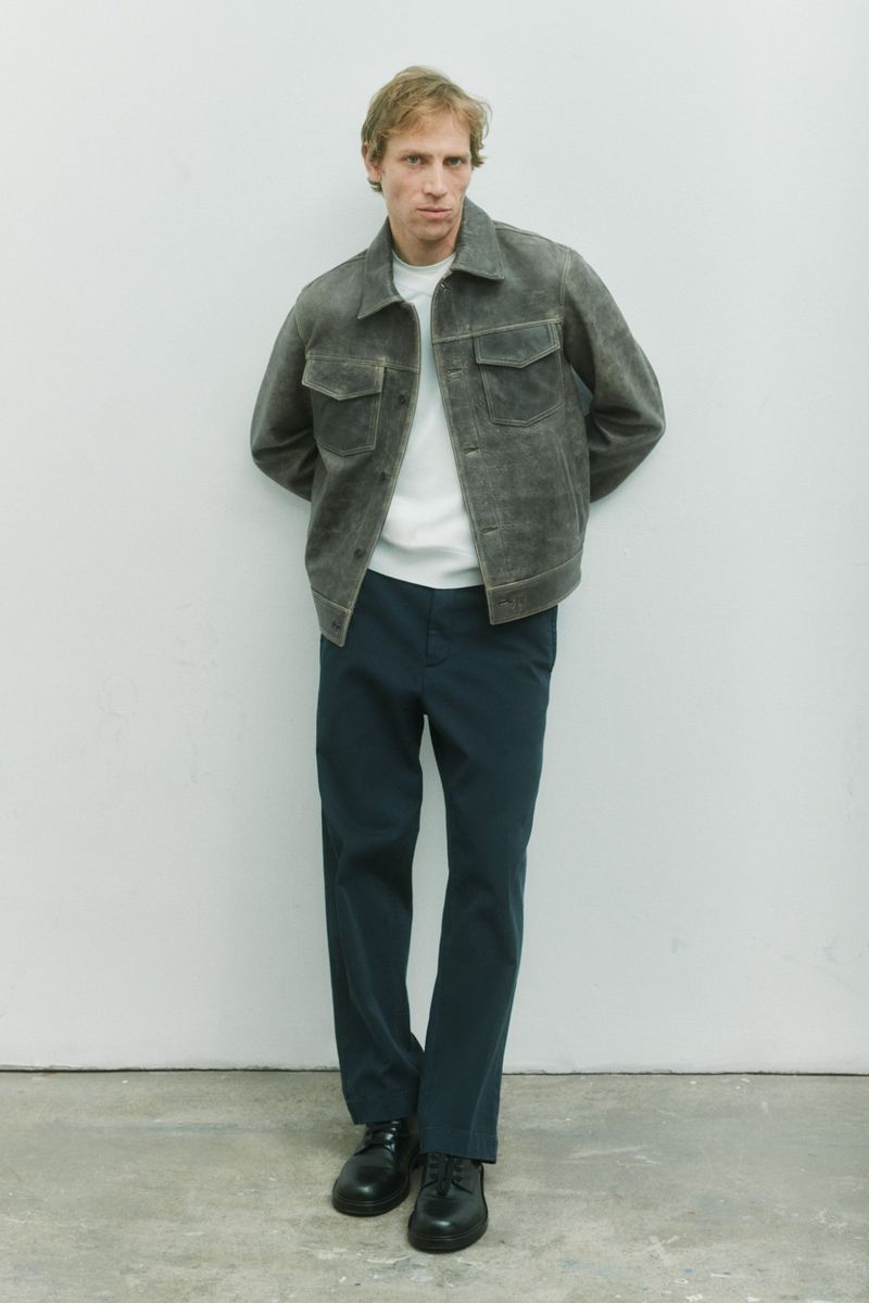Model Mike Vasilakis posing against a pale studio wall in a grey trucker jacket, white shirt, dark trousers, and black shoes.