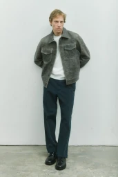 Model Mike Vasilakis posing against a pale studio wall in a grey trucker jacket, white shirt, dark trousers, and black shoes.