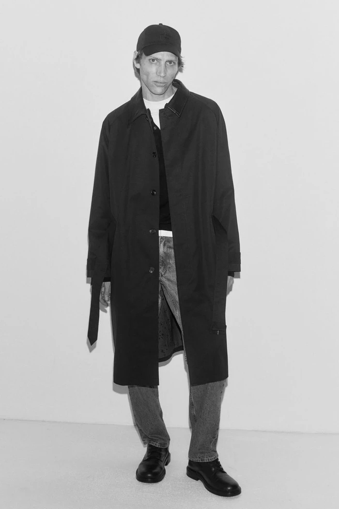 Mike Vasilakis wearing a long black coat, baseball cap, and jeans against a plain white studio backdrop for Rag & Bone.