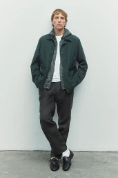 Model Mike Vasilakis standing against a white backdrop wearing a dark green jacket, grey trousers, and black loafers.
