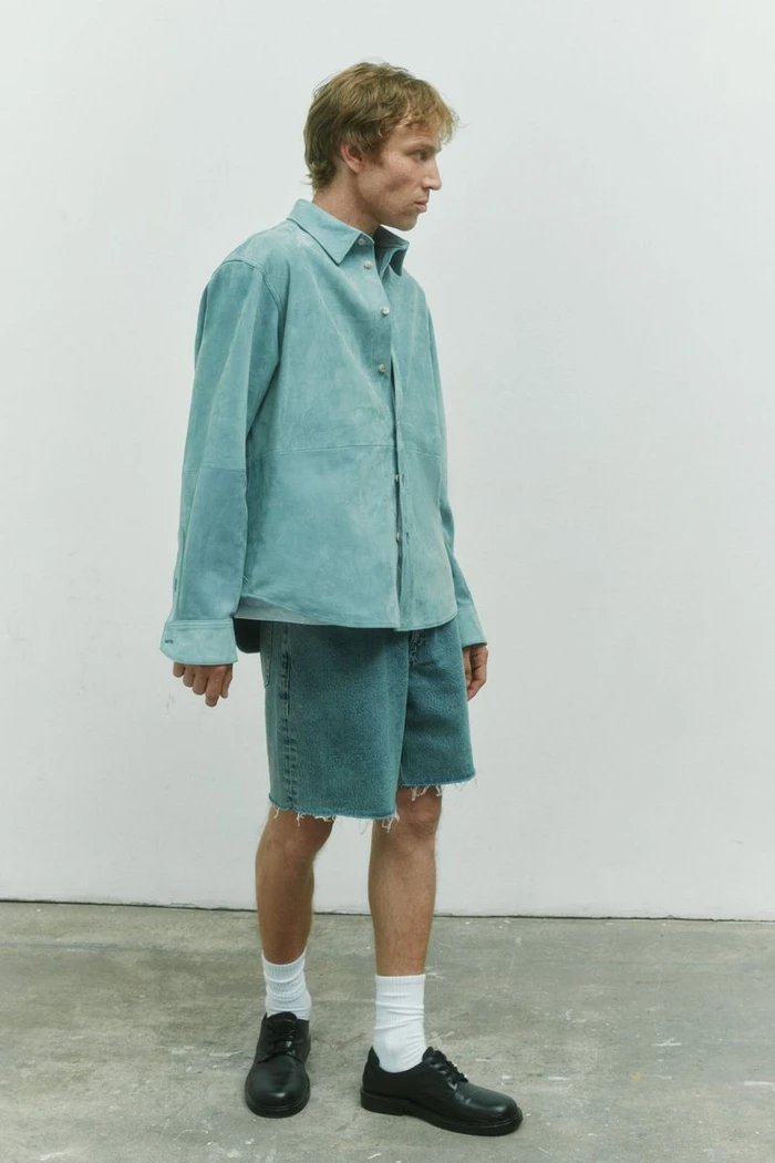 Model Mike Vasilakis standing in profile wearing a pale teal suede overshirt and denim shorts for Rag & Bone menswear.
