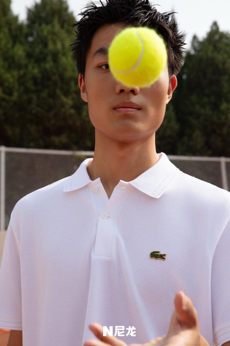 A young man in a white polo shirt poses with a tennis ball in front of his eye on a tennis court with trees in the background.