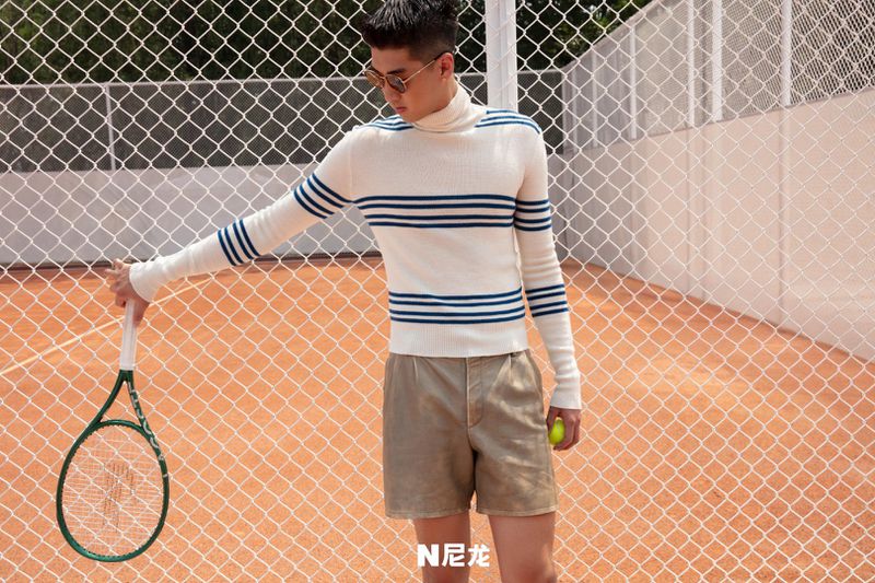 Male model poses on tennis court wearing a striped turtleneck sweater and shorts, holding a racquet and a tennis ball.