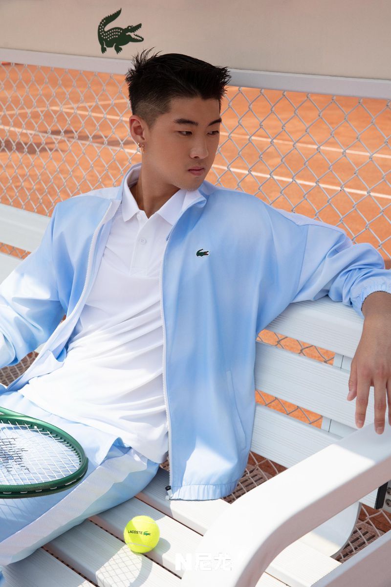 Stylish Asian man in a Lacoste tennis outfit poses on a court bench with a racket and ball, illuminated by natural light.