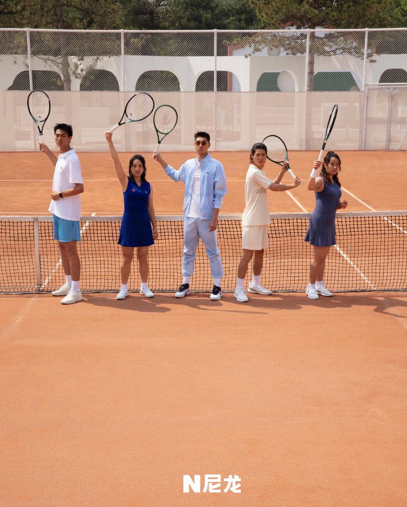 Five young adults stand on a tennis court holding rackets, posing in sportswear against a building with arches and outdoor setting.