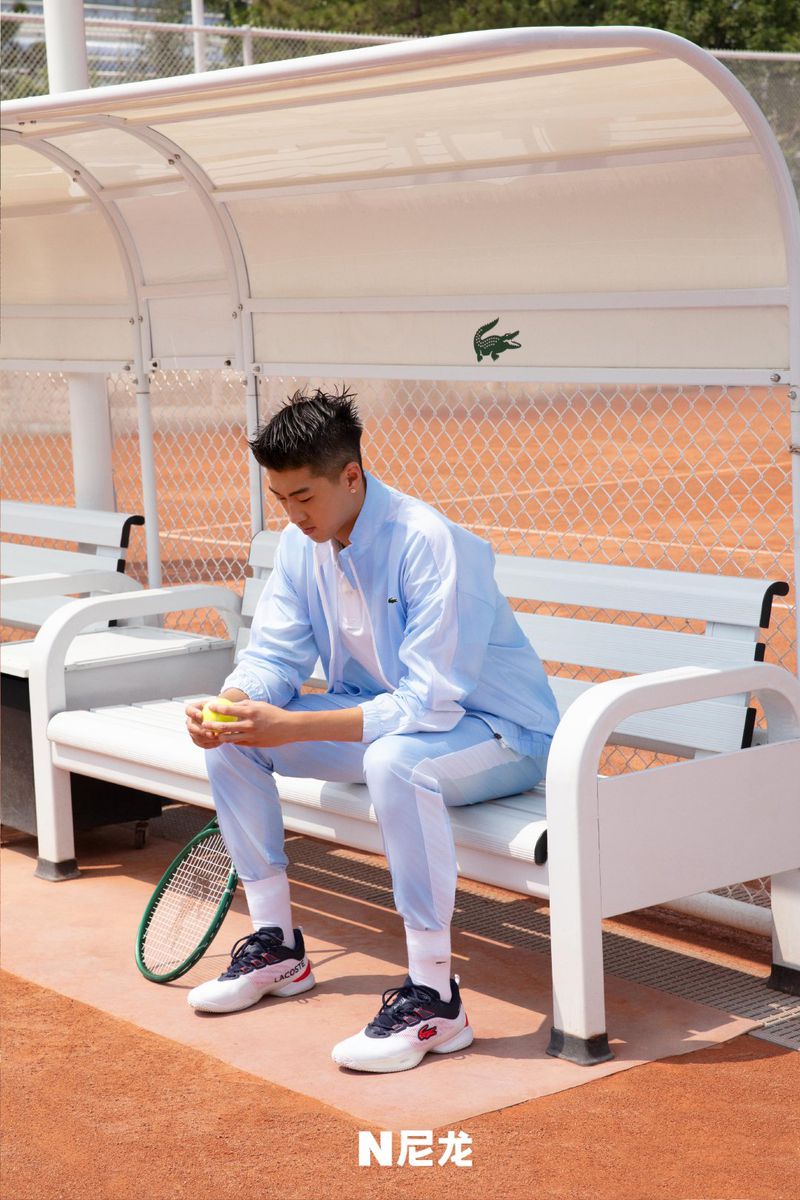 Male tennis player in light blue tracksuit sitting on court bench holding tennis ball on sunny day, Lacoste logo in background.