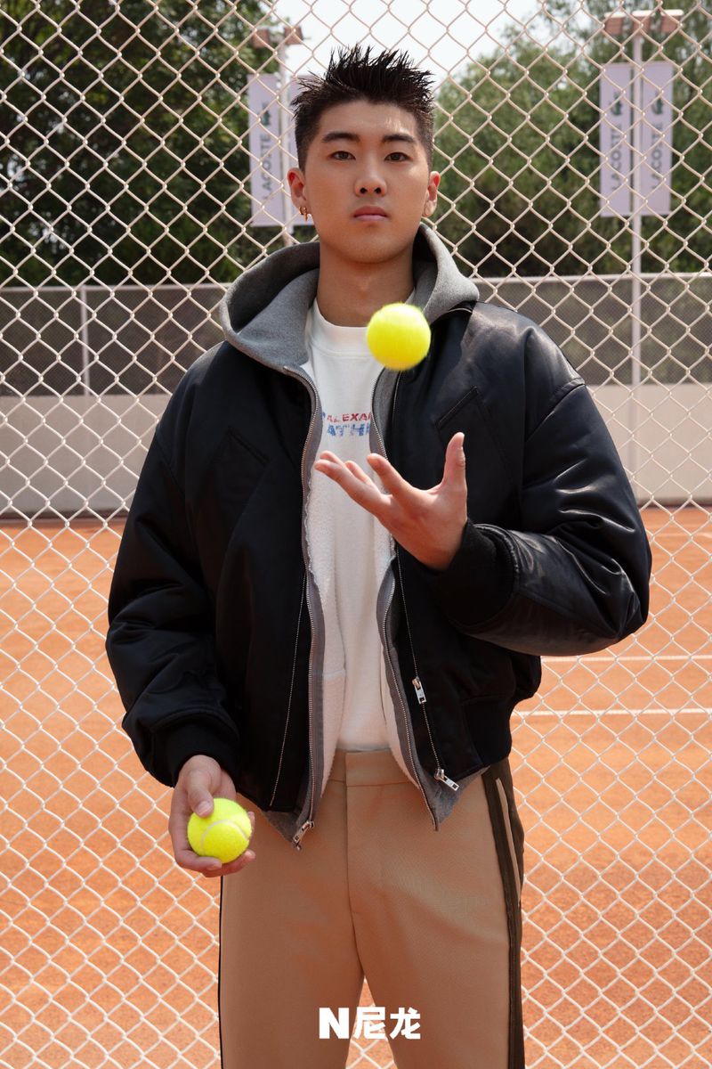 A young man with short dark hair is juggling a tennis ball in front of a tennis court while holding another ball in his hand.