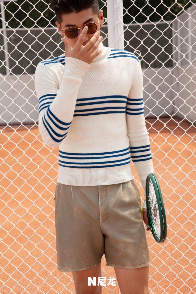 Stylish man in striped sweater and tan shorts poses with tennis racket on an orange court, wearing sunglasses and holding hand to face.