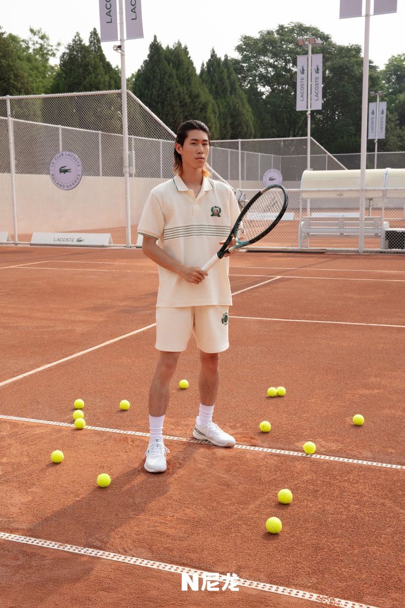 Male tennis player in cream shorts and shirt holds a tennis racket on a clay court with tennis balls scattered around, outdoor setting.