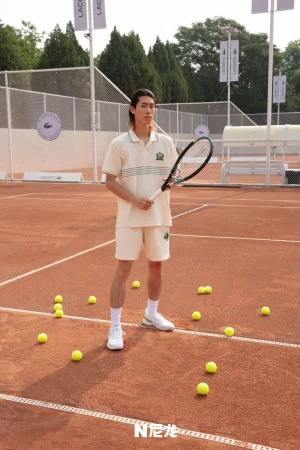Male tennis player in cream shorts and shirt holds a tennis racket on a clay court with tennis balls scattered around, outdoor setting.