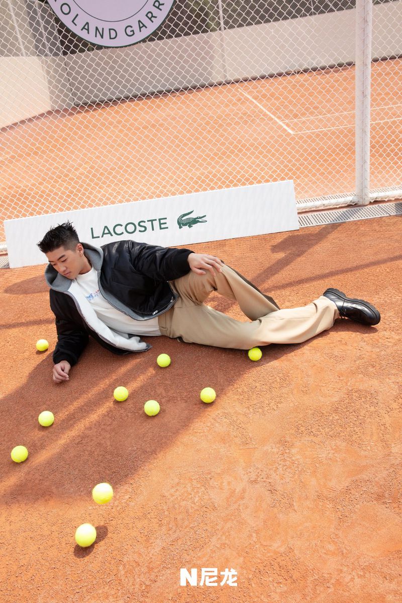 Asian man in jacket poses on clay surface surrounded by tennis balls with Lacoste signage and tennis net in the background.