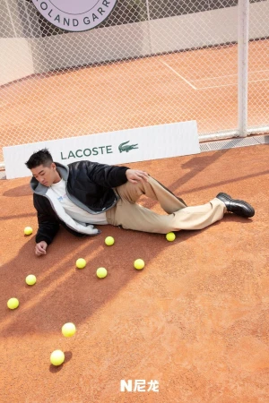 Asian man in jacket poses on clay surface surrounded by tennis balls with Lacoste signage and tennis net in the background.
