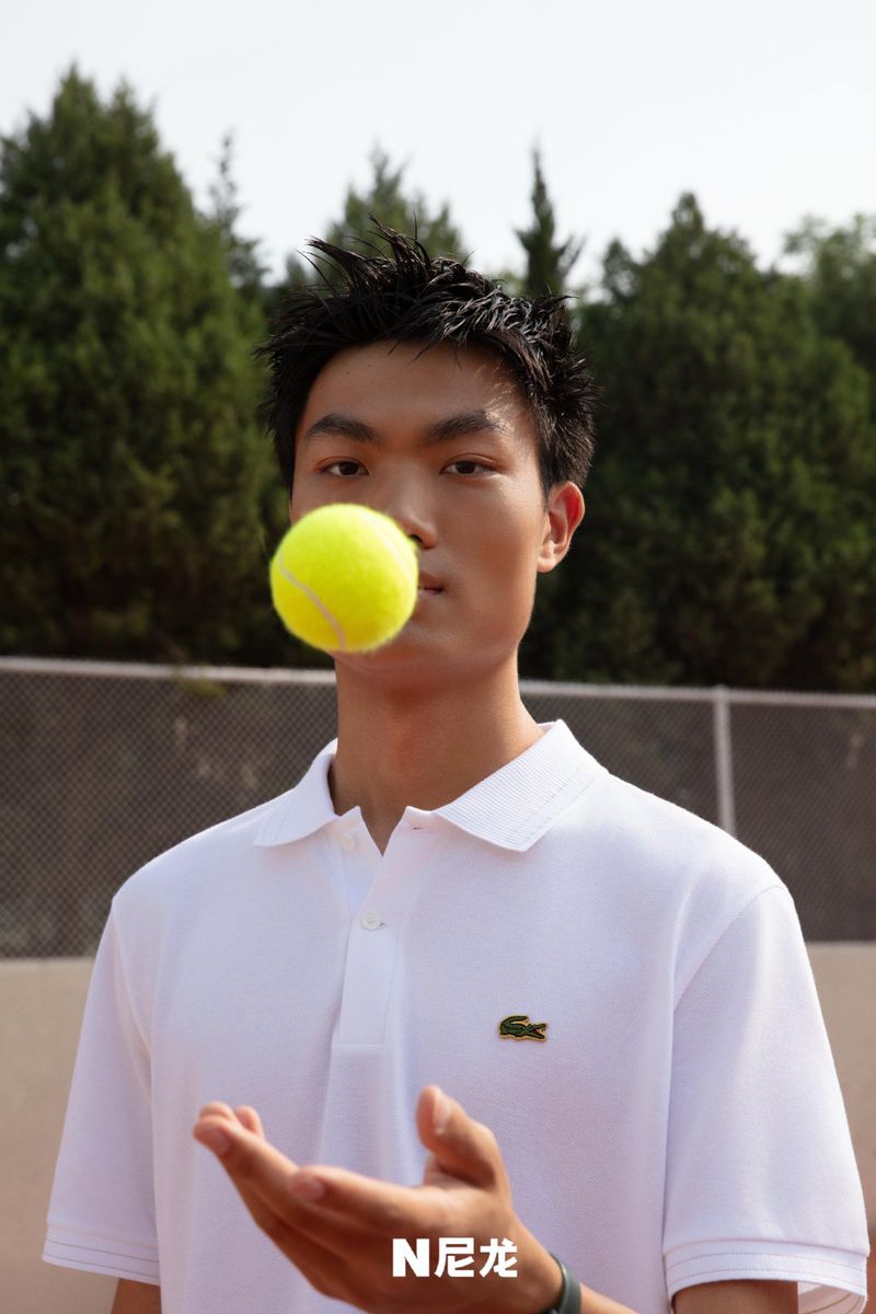 Young Asian man balances a tennis ball on his mouth, wearing a white polo shirt on a tennis court, with green trees in background.