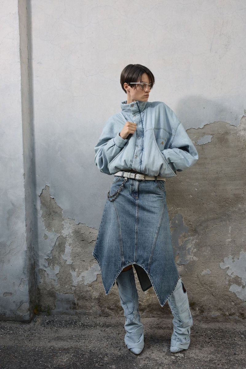 Model Tanya Churbanova wearing a light denim jacket, asymmetrical denim skirt, and denim boots against a weathered concrete wall.